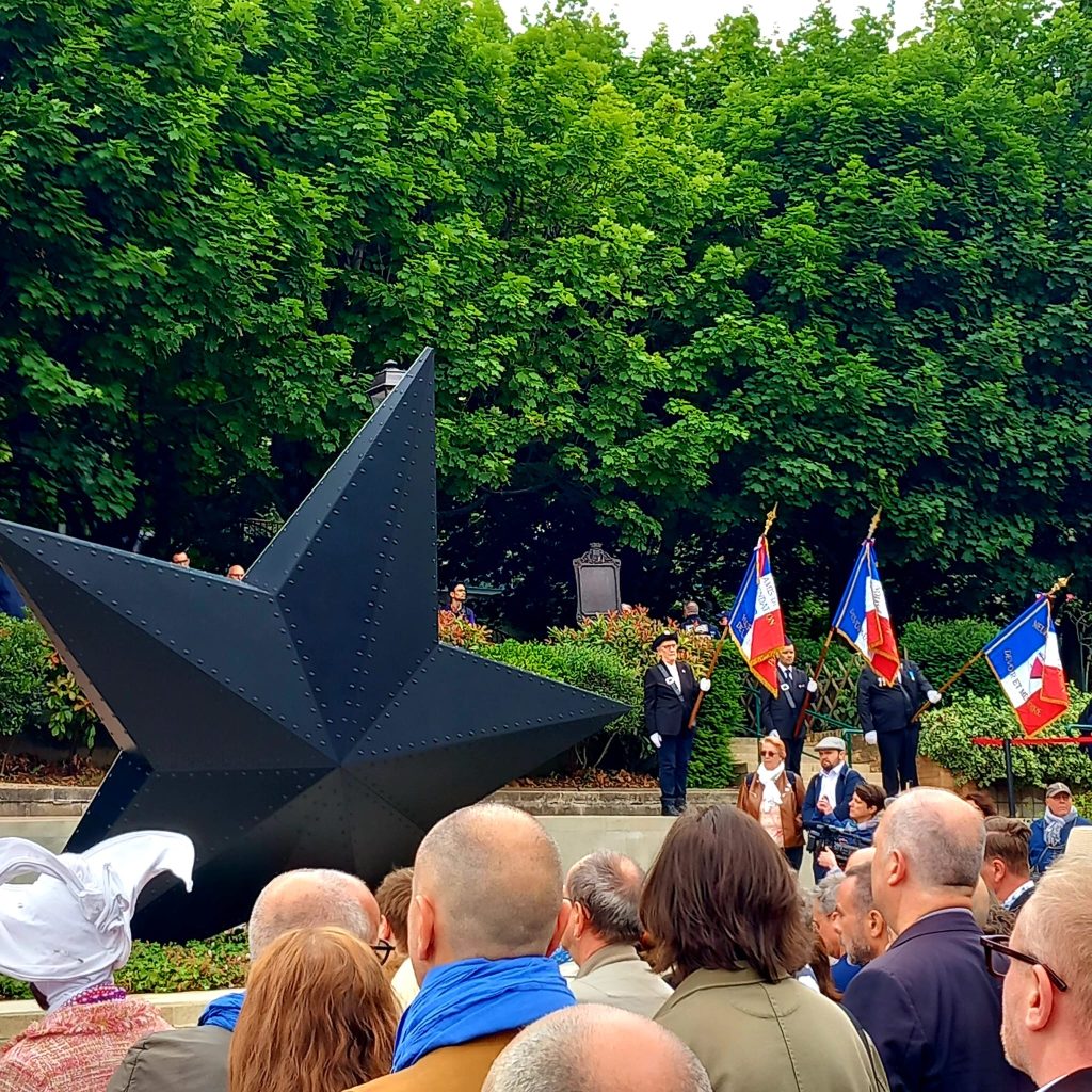 monument, foule au premier plan, verdure en second plan. 3 porte drapeau sur la droite